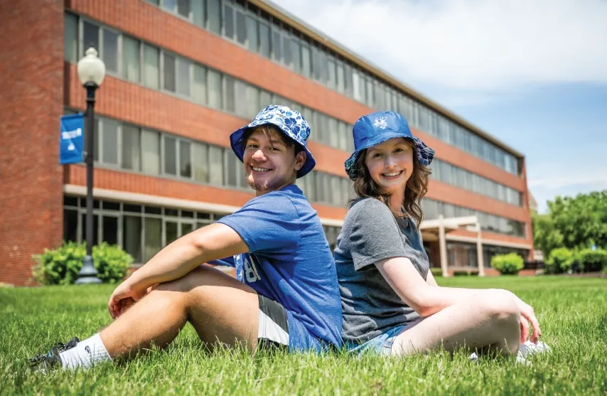 Two students wearing bucket hats smiling and sitting back to back.