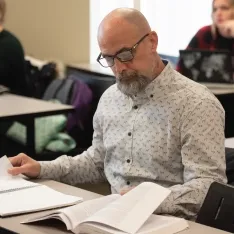 A man with glasses and a beard sitting at a desk with books and papers.